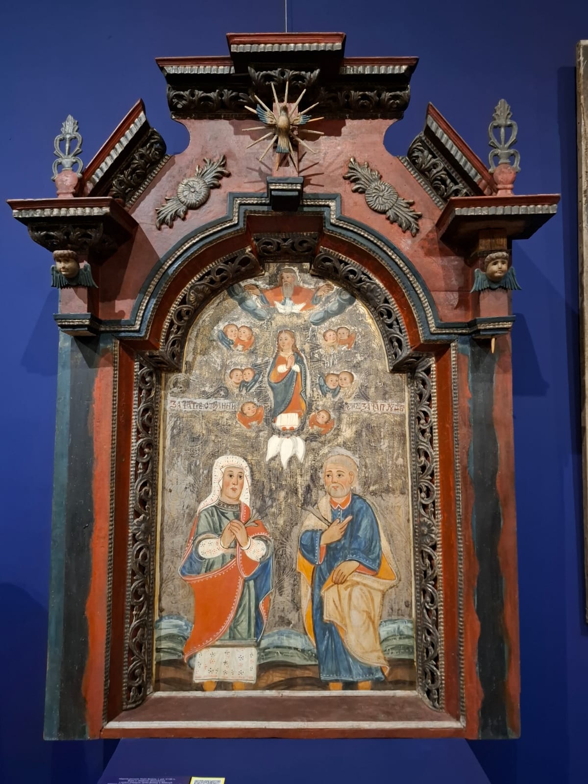 A polychrome religious icon in a richly carved frame depicting a Marian scene with figures of saints and angels.