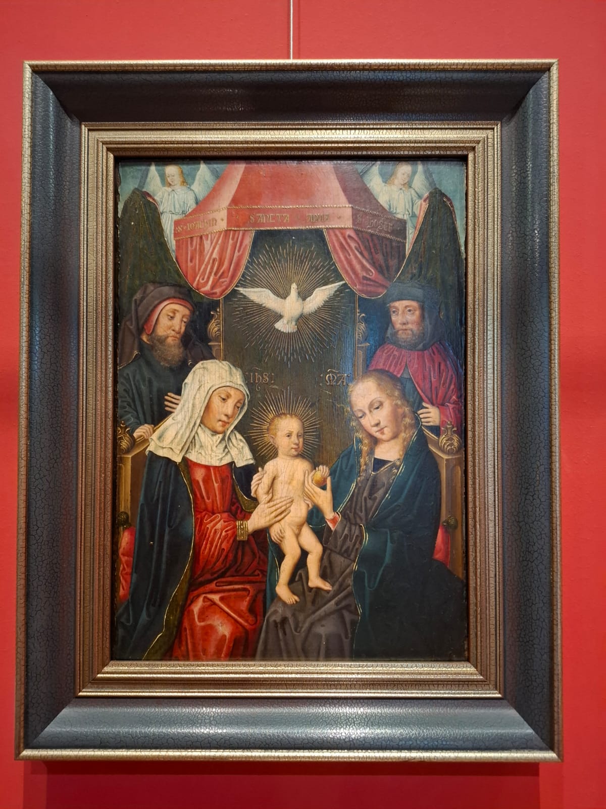 A religious painting depicting the Baby Jesus held by Mary and Saint Anne, with angels and the dove of the Holy Spirit above the scene.