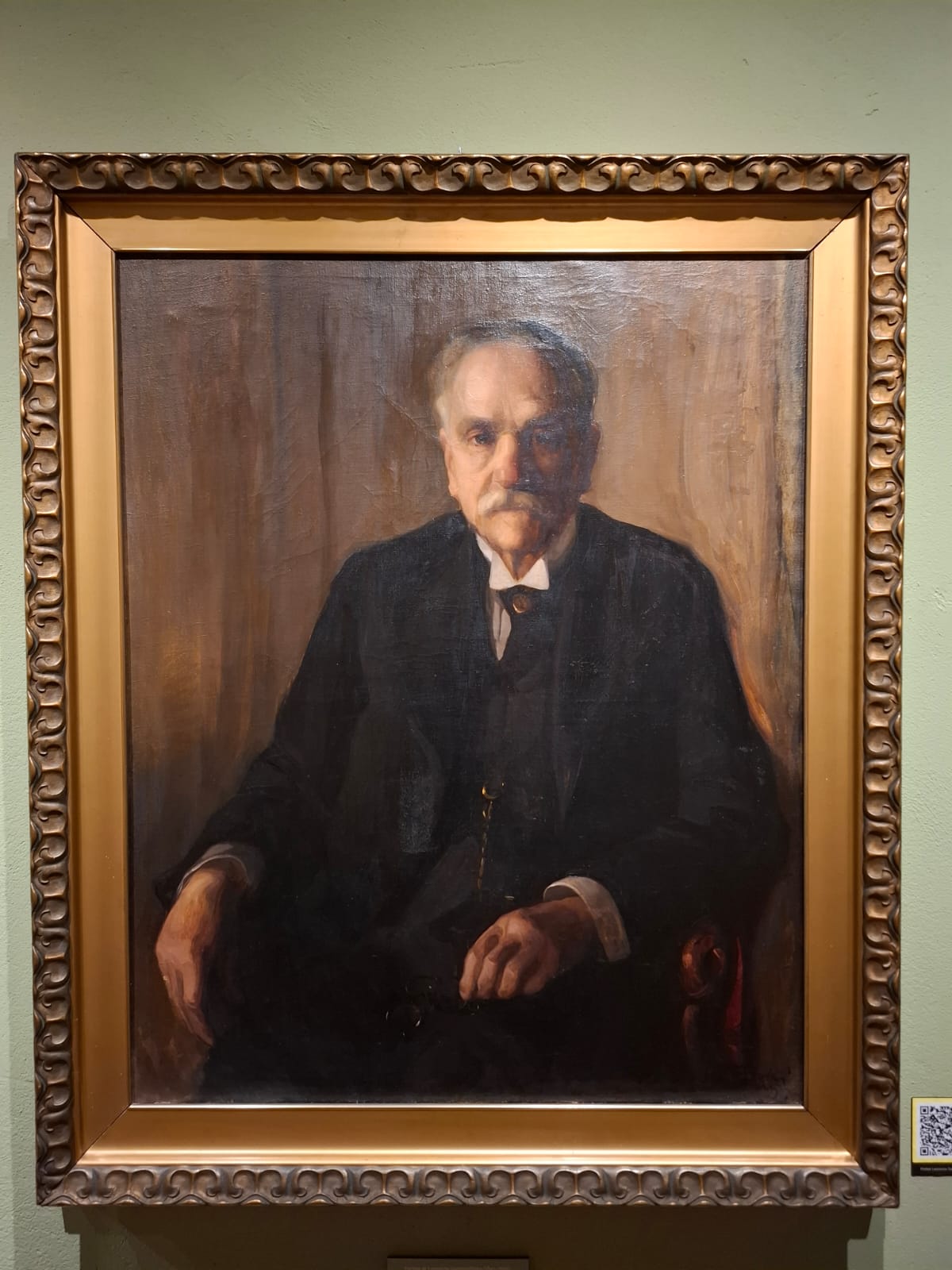 Portret starszego mężczyzny w ciemnym stroju, siedzącego w fotelu, namalowany w realistycznym stylu i oprawiony w ozdobną ramę.